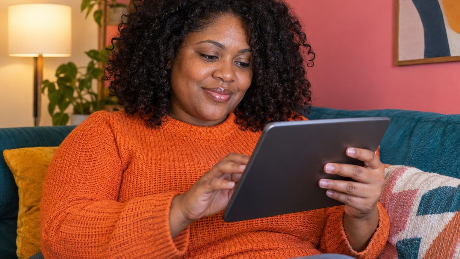 Woman with natural curls in an orange sweater reviews a tablet on a colourful sofa.
