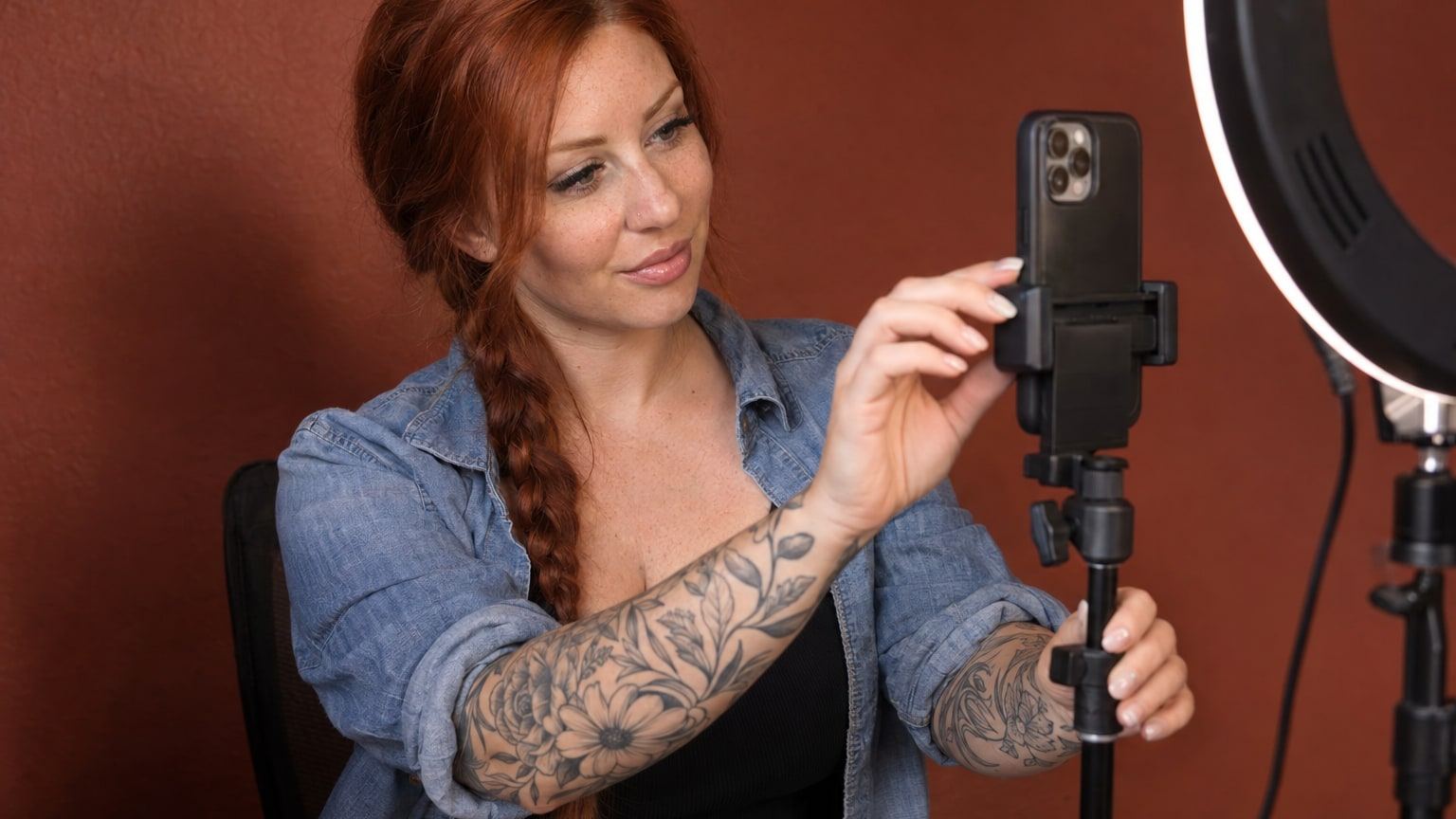 Woman with a red braid and tattooed arms adjusts her phone beside a ring light.